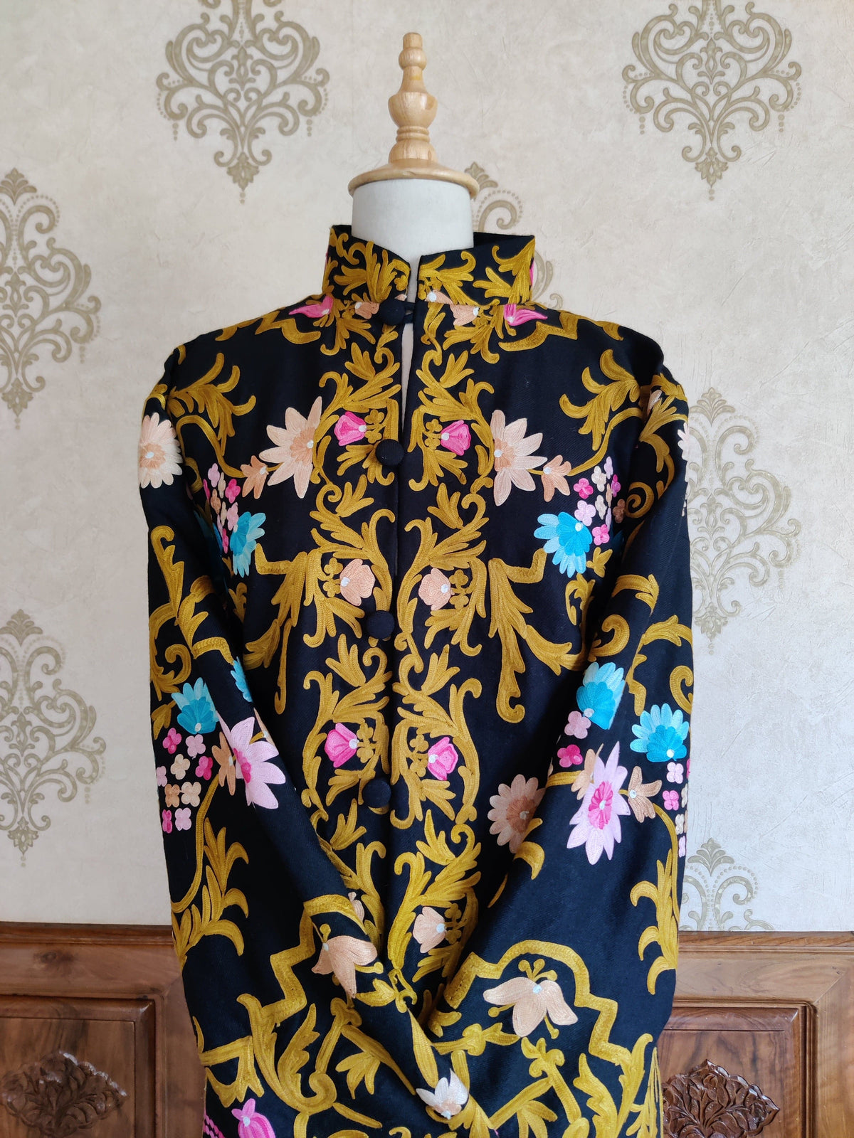 Front detail of a black coat with one sleeve across the body, showing golden Aari scrolls and colourful floral embroidery