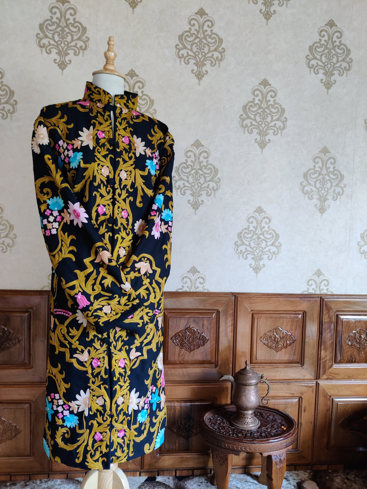 Full front of a long black coat with golden Aari vines, pink and blue flowers and one sleeve folded across the waist