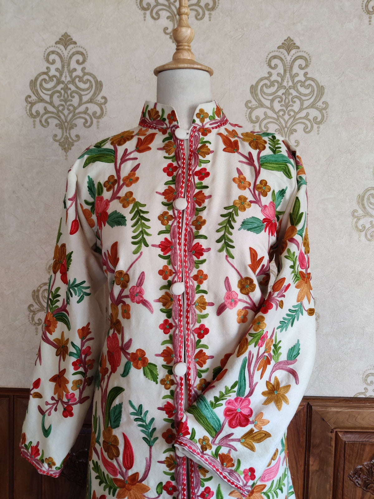 Upper front of a white Aari jacket with colourful flowers, green leaves, a stand collar and one sleeve across the waist