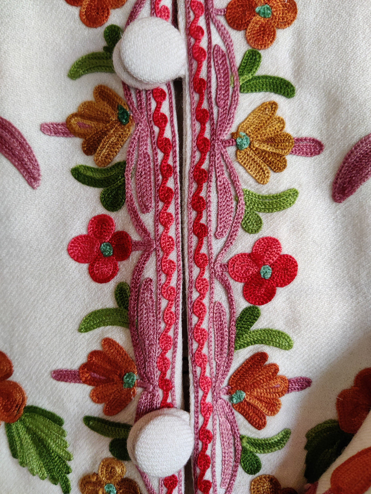 Close-up of white covered buttons and pink placket edging bordered by red and ochre Aari flowers on a white jacket
