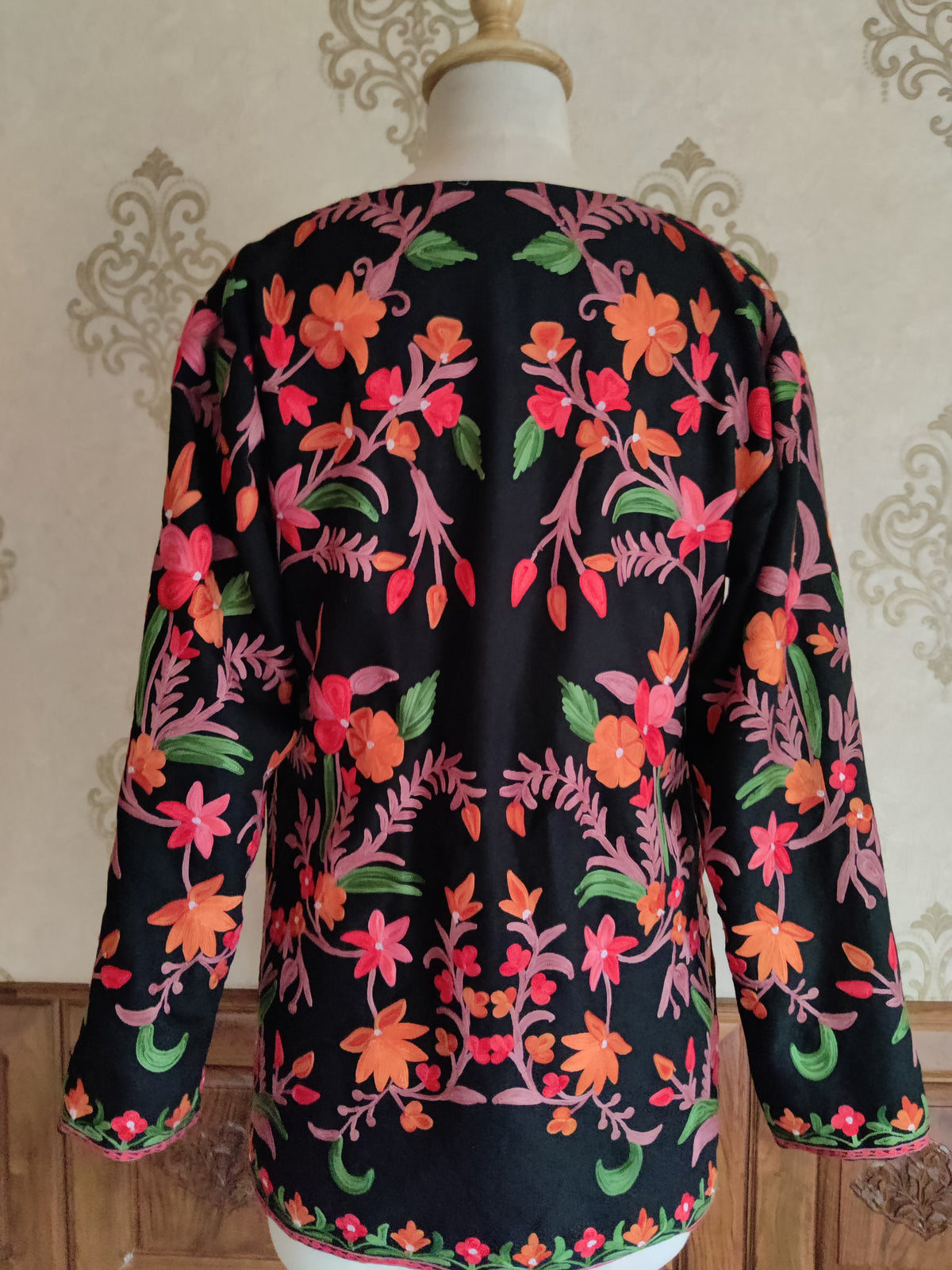 Back of a black short jacket with symmetrical pink and orange Aari flower sprays, green leaves and embroidered cuffs