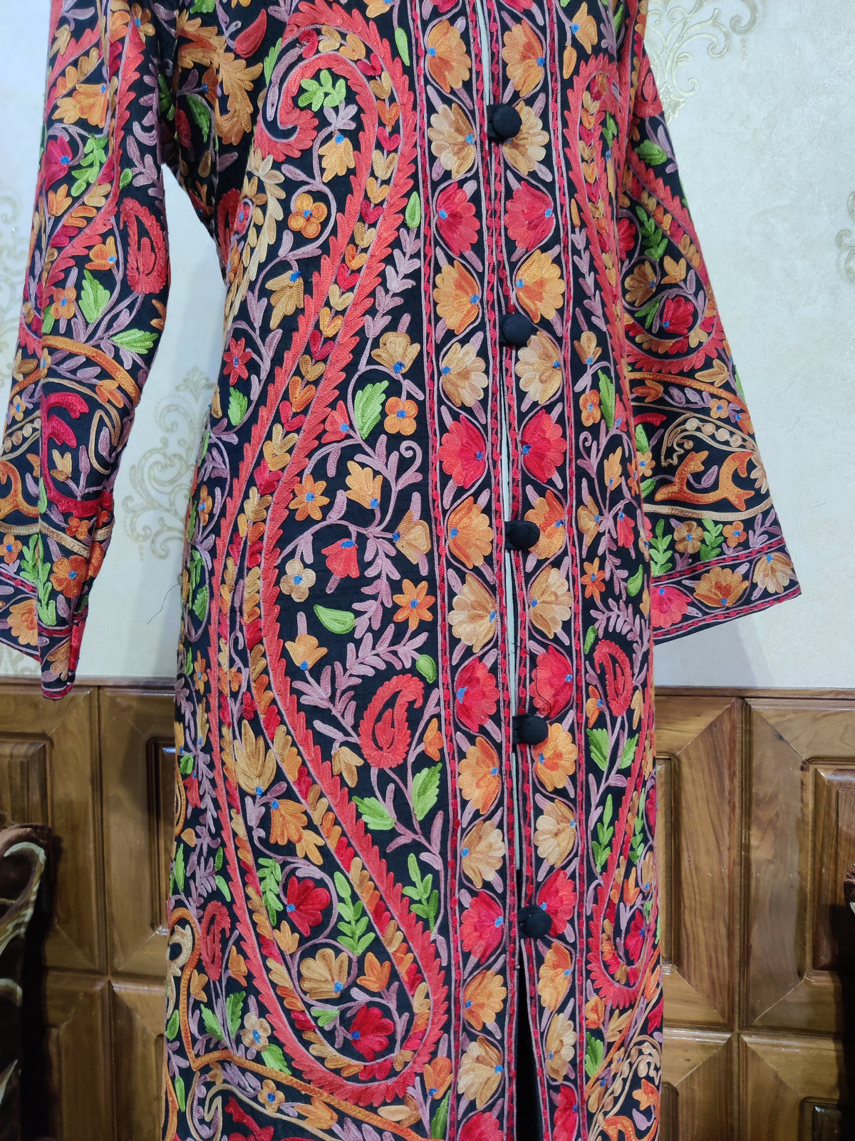 Closer front of the black jacket, showing sweeping multicoloured Aari paisleys beside the flower-bordered button opening.