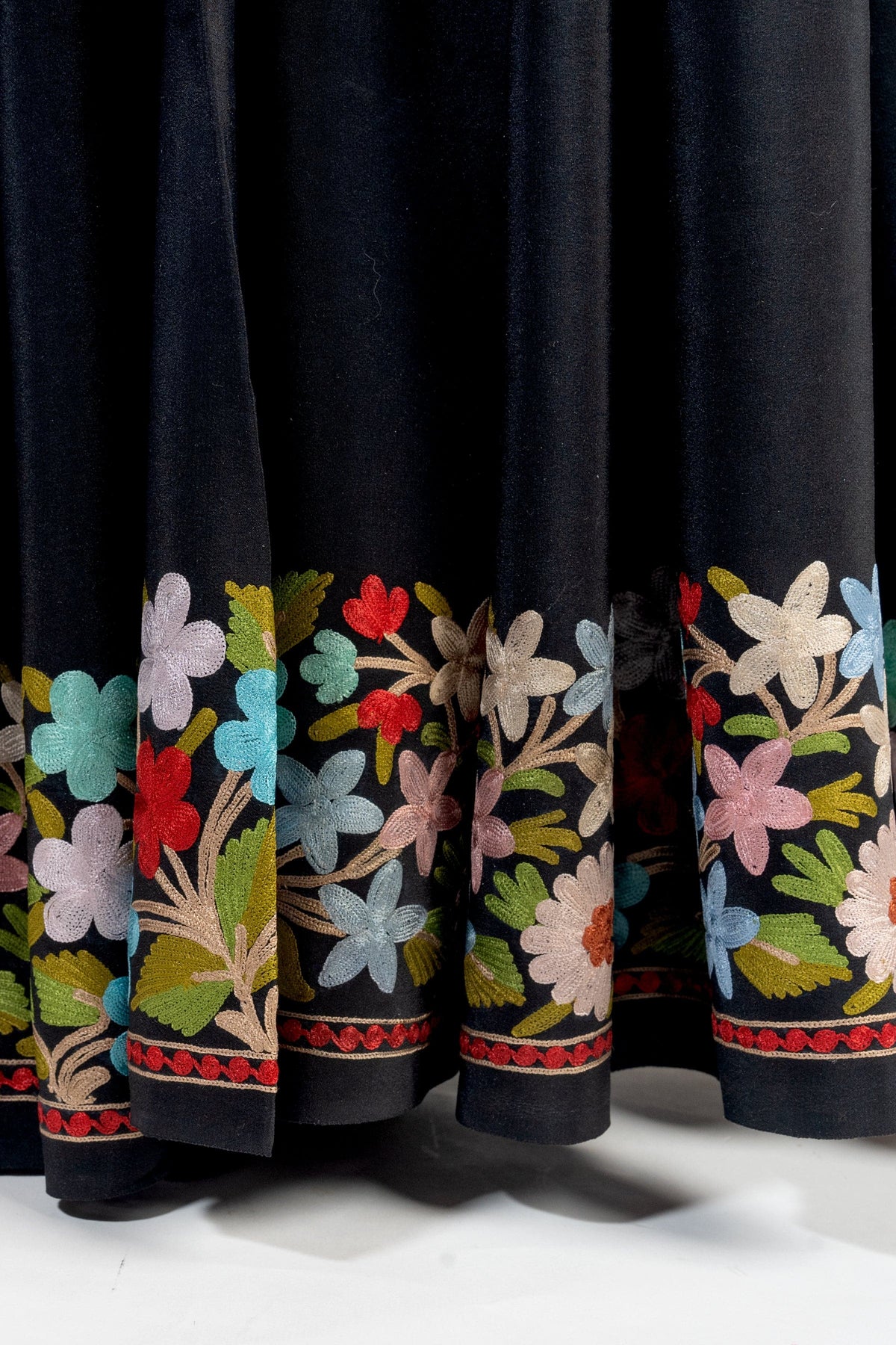 Close-up of pleated black saree fabric with multicoloured Aari flowers and green leaves above narrow red edging