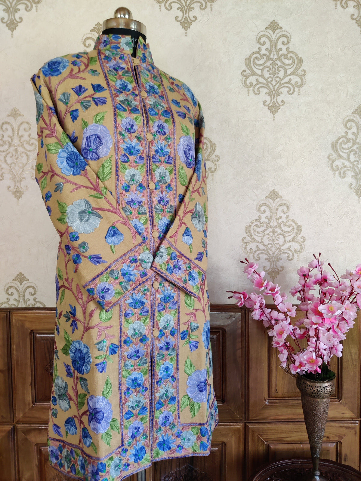 Angled full view of a golden-beige floral-embroidered jacket with crossed sleeves, a band collar and fabric-covered buttons.