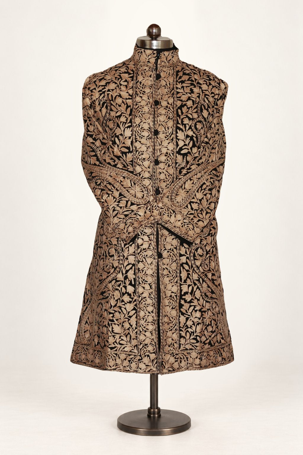 Front of a black wool jacket on a mannequin, densely covered with gold-toned Aari paisleys and leafy vines, with sleeves crossed.