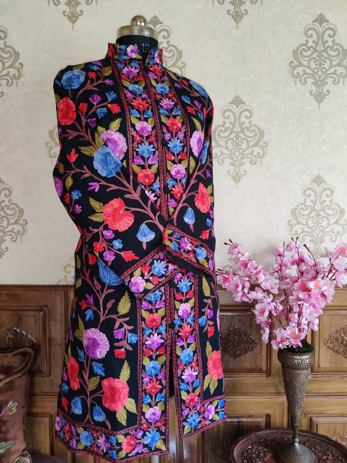Angled full view of a black jacket on a mannequin with crossed sleeves, colourful floral embroidery and a stand collar.