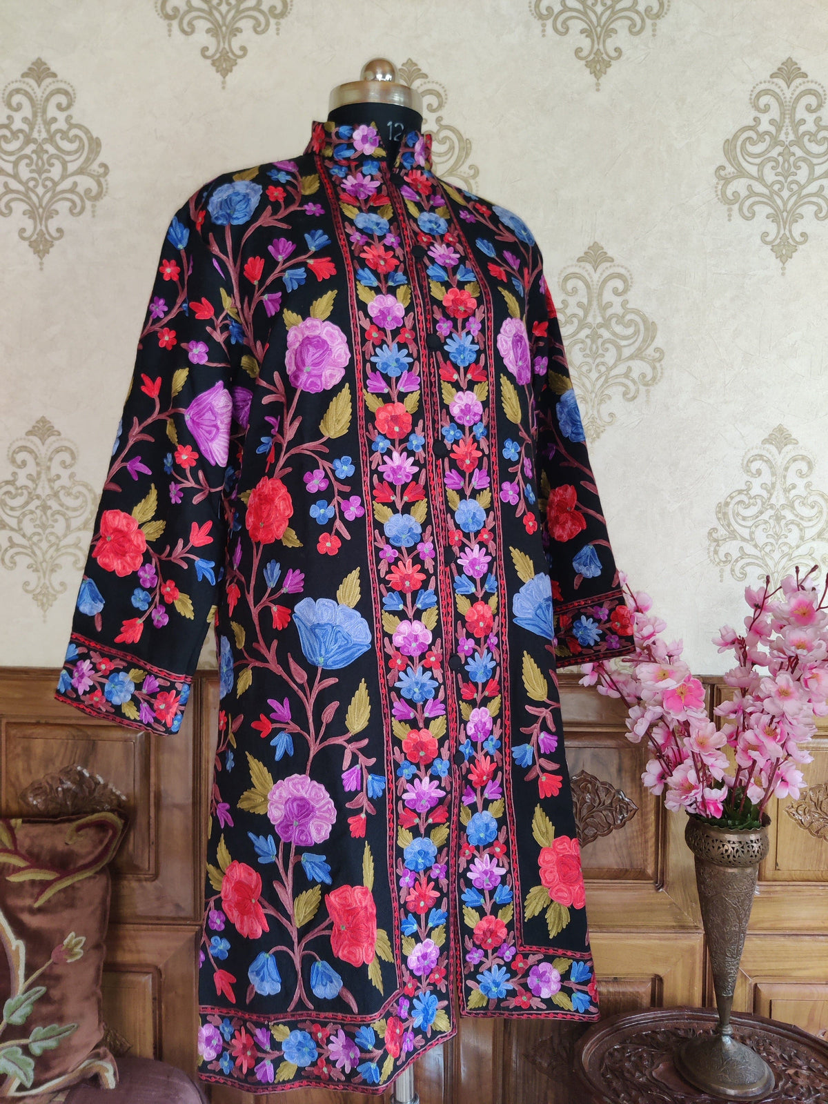 Front view of a long black jacket with sleeves lowered, showing colourful flowers and embroidered borders along the opening, cuffs and hem.
