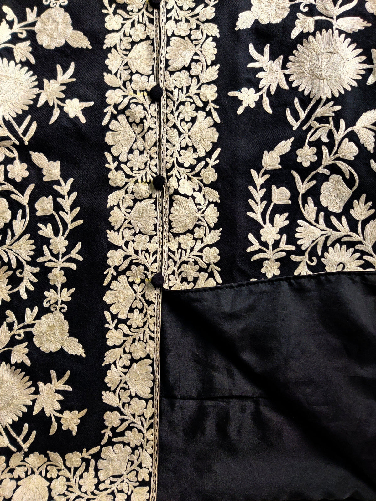 Black jacket laid flat with one edge turned back to show the plain lining beside white floral embroidery and covered buttons