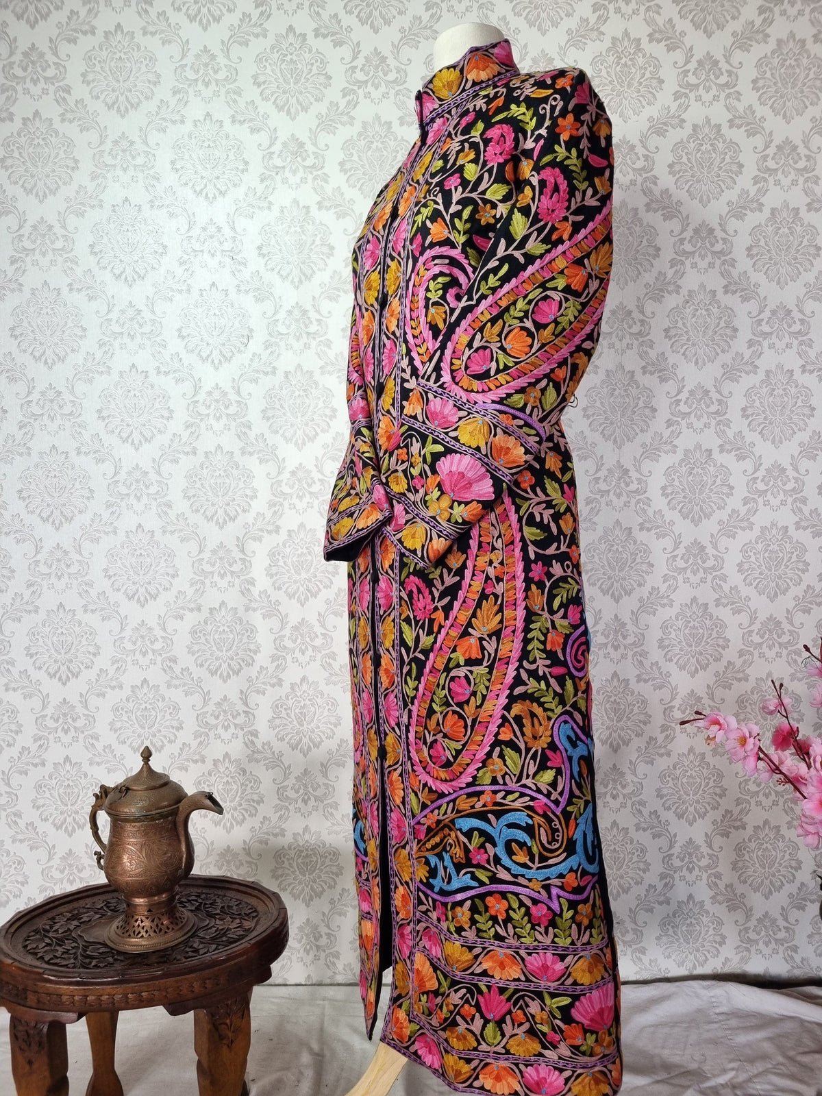 Angled full-length view of the black jacket with crossed sleeves, bright floral vines and a patterned hem.