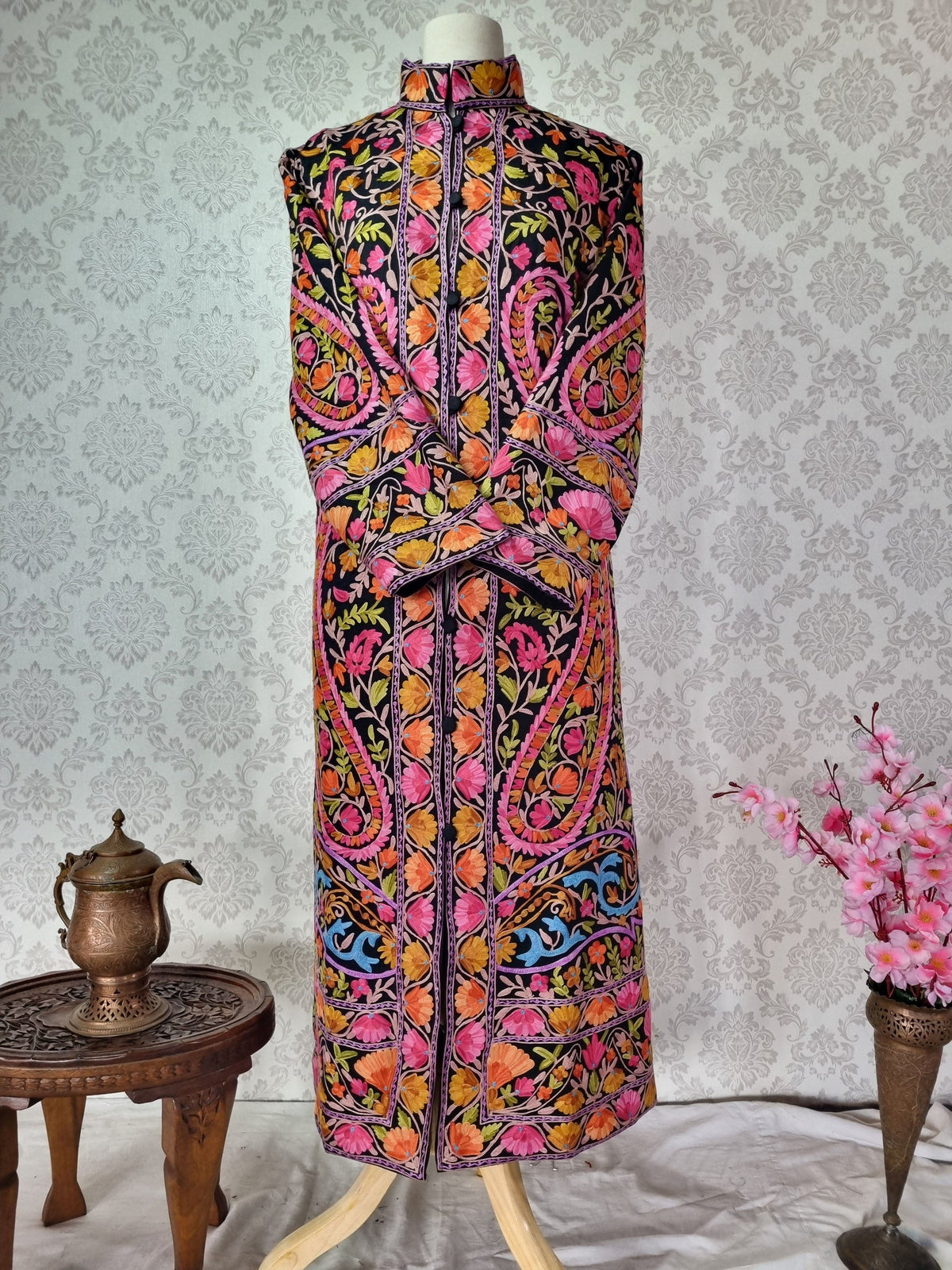 Long black wool jacket on a mannequin with crossed sleeves, dense colourful floral embroidery and oversized paisley outlines.