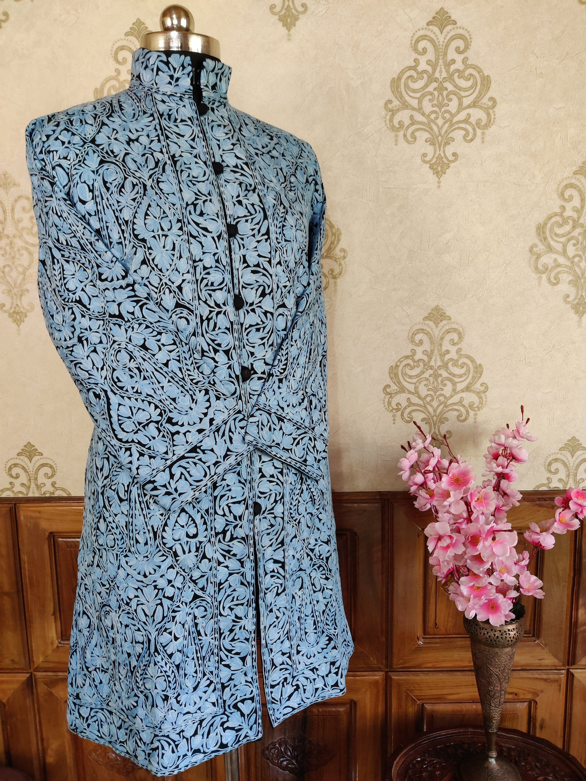 Angled full front of a black long jacket with dense pale blue Aari paisley embroidery, a stand collar and crossed sleeves