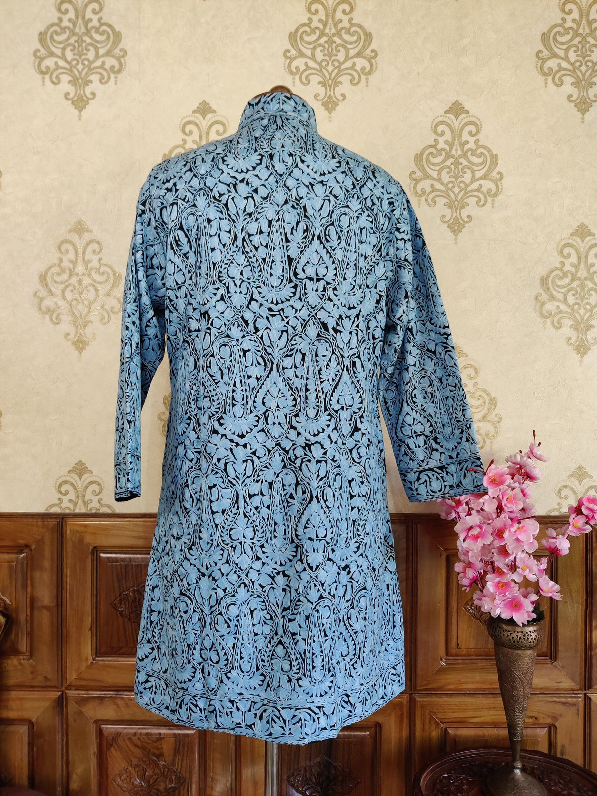 Back of a black long jacket covered with symmetrical pale blue Aari paisley and floral embroidery and a narrow patterned hem