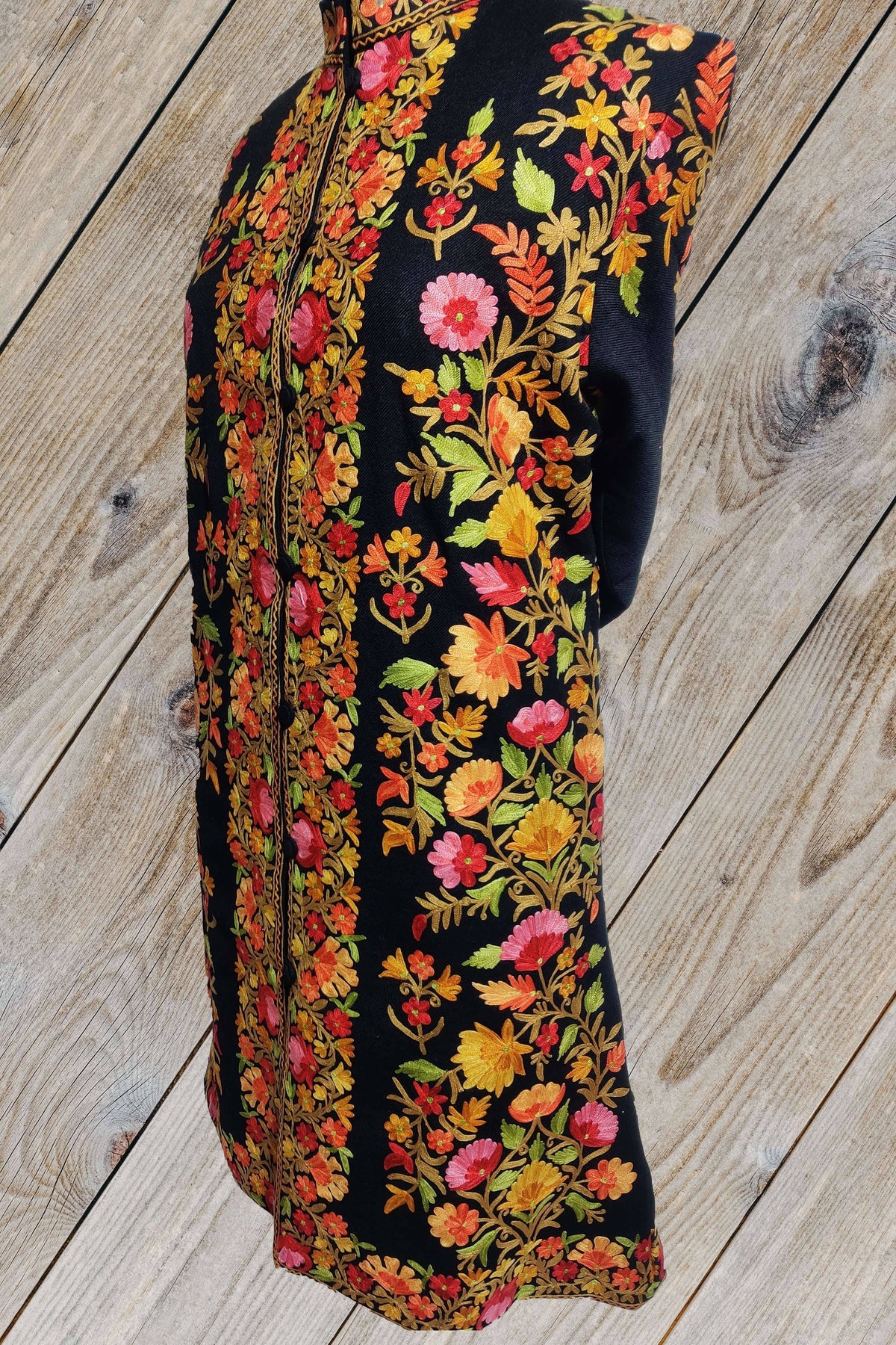 Three-quarter front of a black long jacket showing pink, orange and yellow Aari flowers with green leaves along the front and side