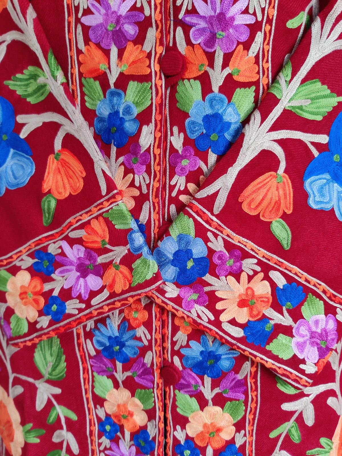 Close-up of the maroon jacket's crossed cuffs and buttoned front, with bright multicoloured Aari flowers and green leaves.
