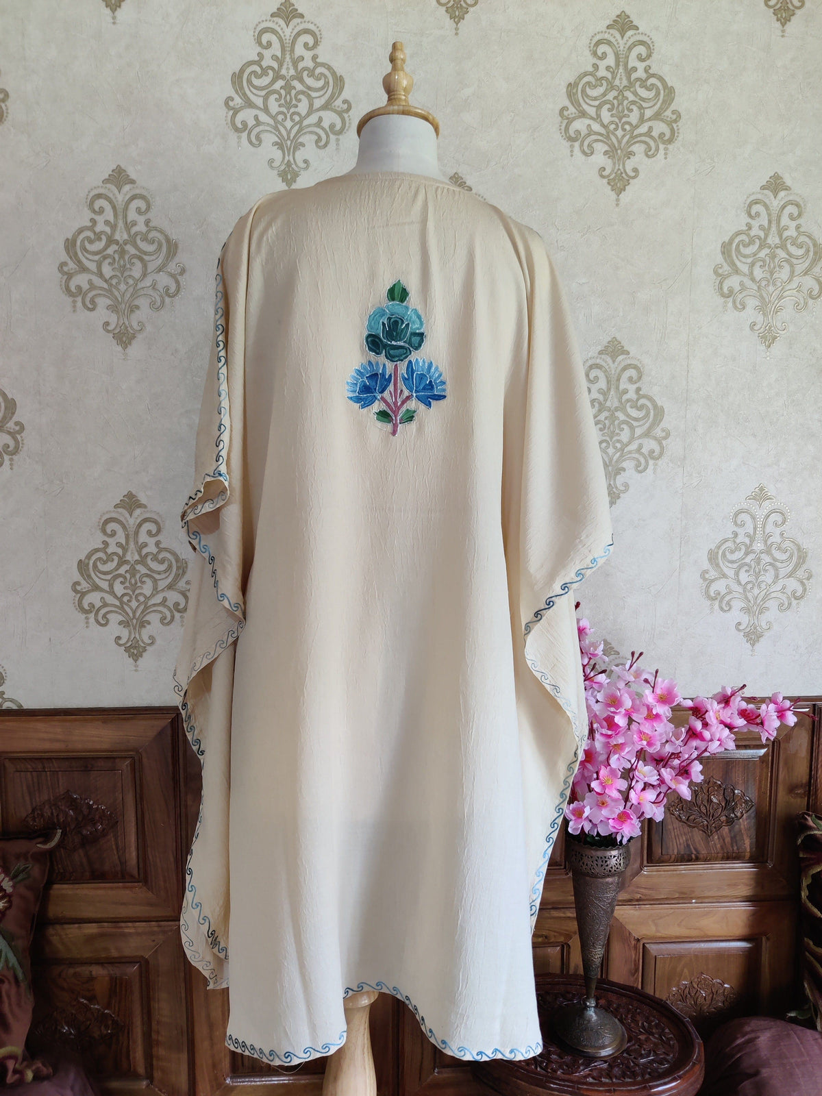 Back of a cream kaftan on a mannequin, with a small blue-and-teal Aari floral motif between the shoulders and blue-edged wide sleeves.