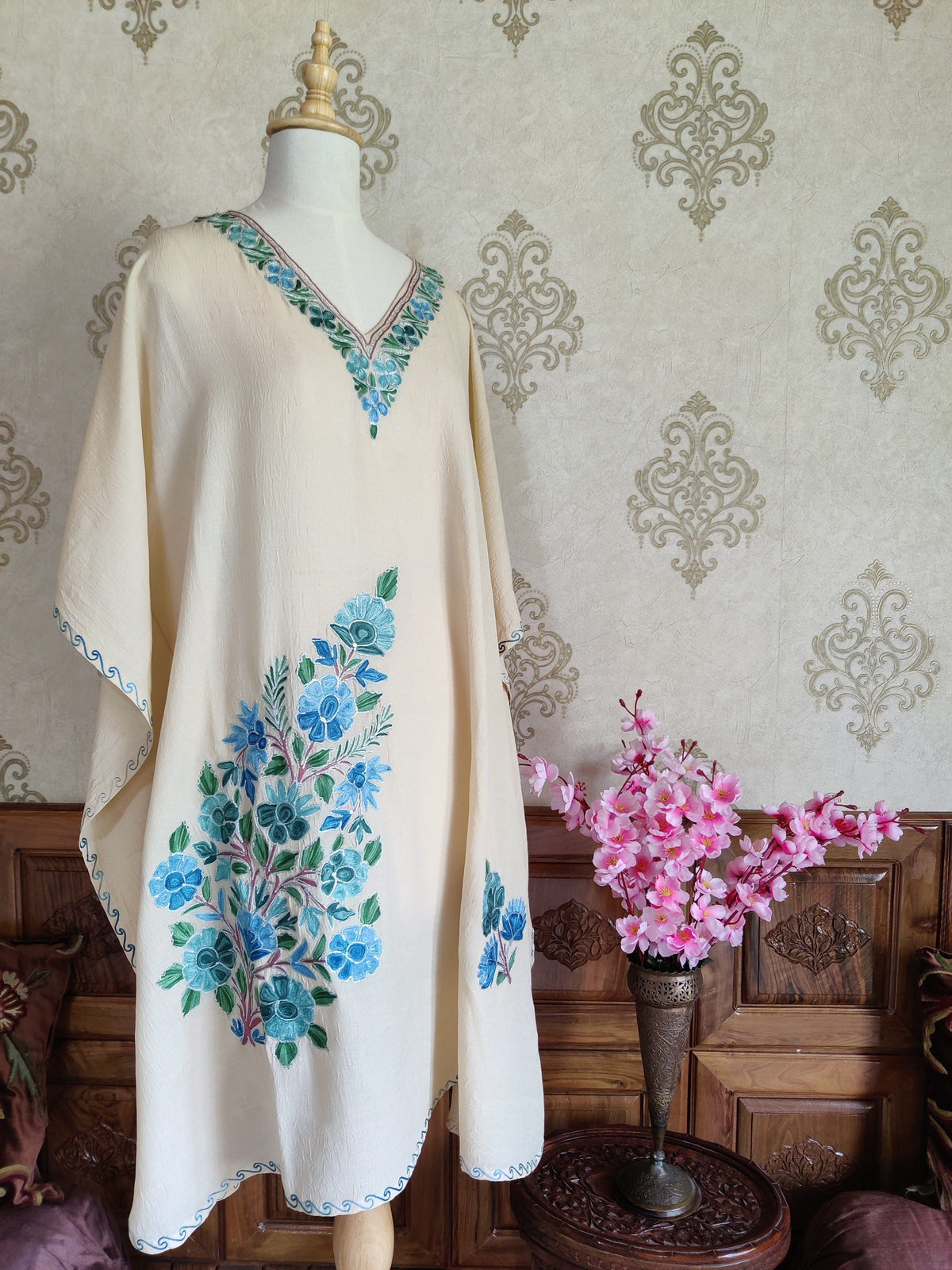 Angled front of a cream kaftan, with a floral V-neck, blue-green Aari flower sprays and wide sleeves edged in blue scrolling stitches.
