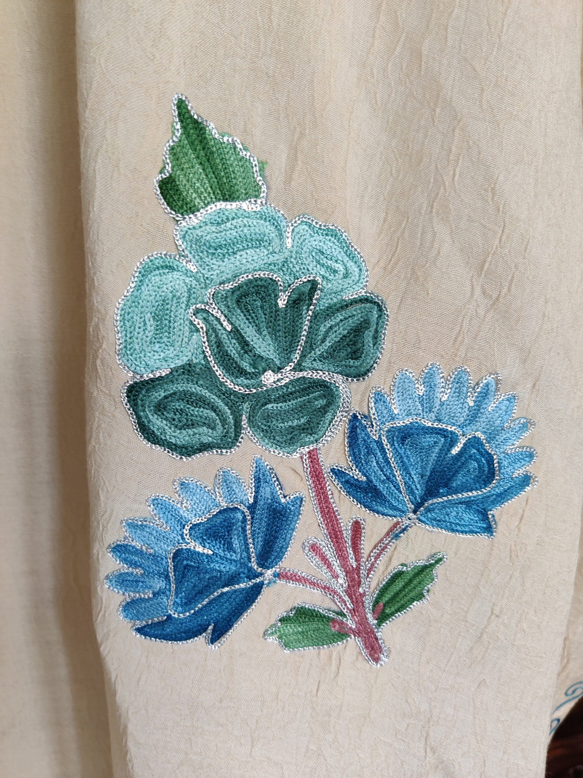 Close-up of a teal rose and two blue Aari flowers with green leaves and pink stems on cream kaftan fabric.