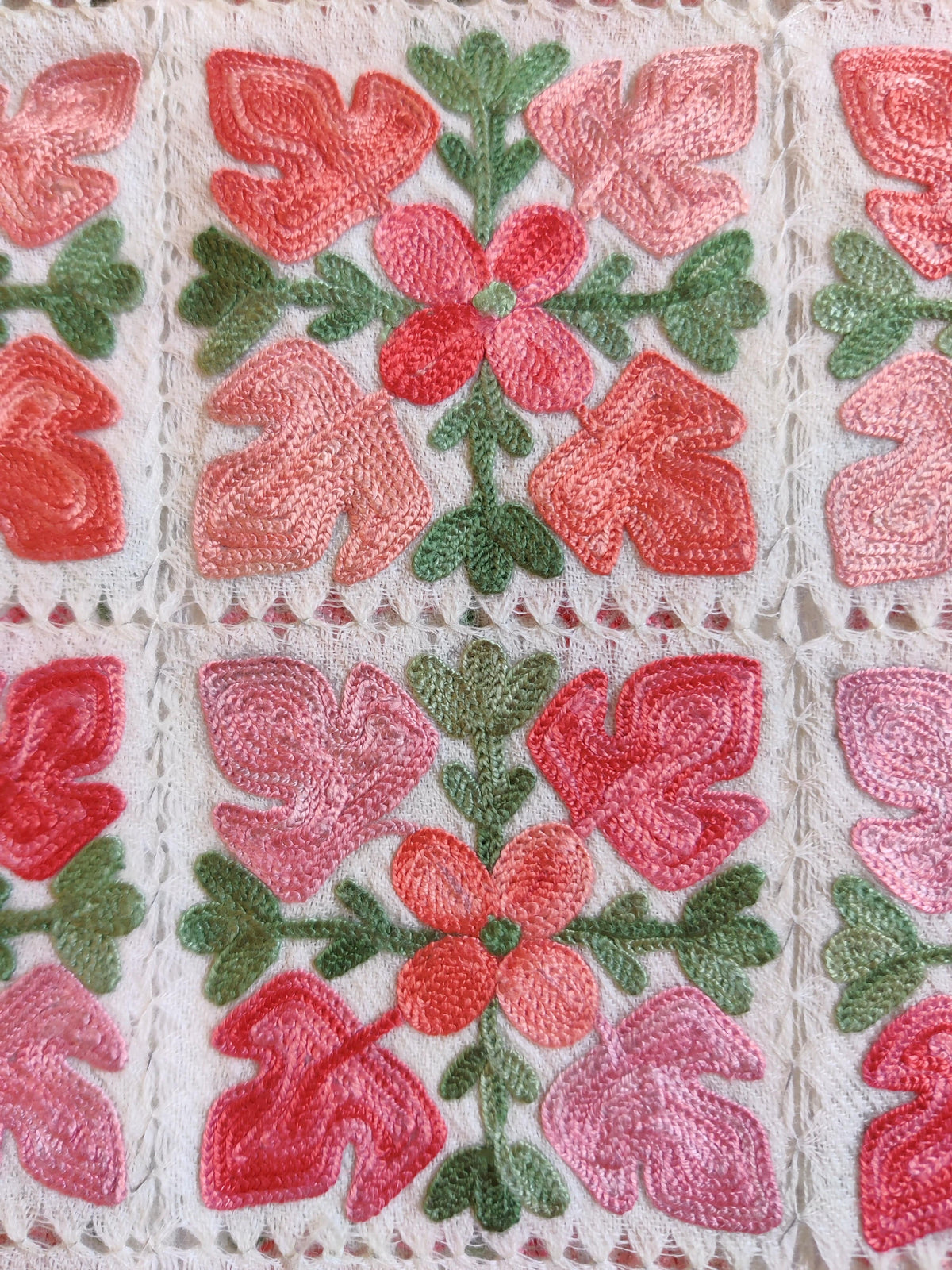 Macro detail of coral-pink flowers and green leaves surrounded by white cutwork stitching.