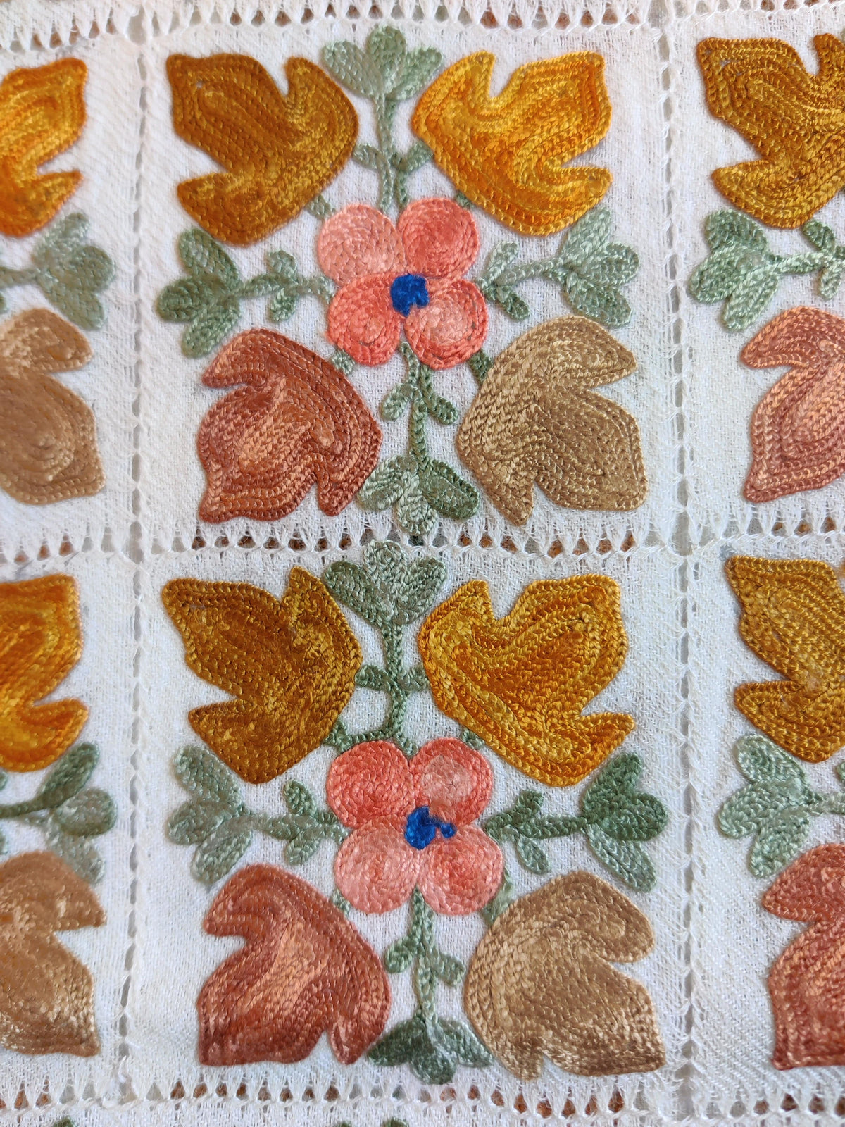 Close-up of peach flowers with blue centres and gold, rust and beige Aari leaves inside white cutwork squares