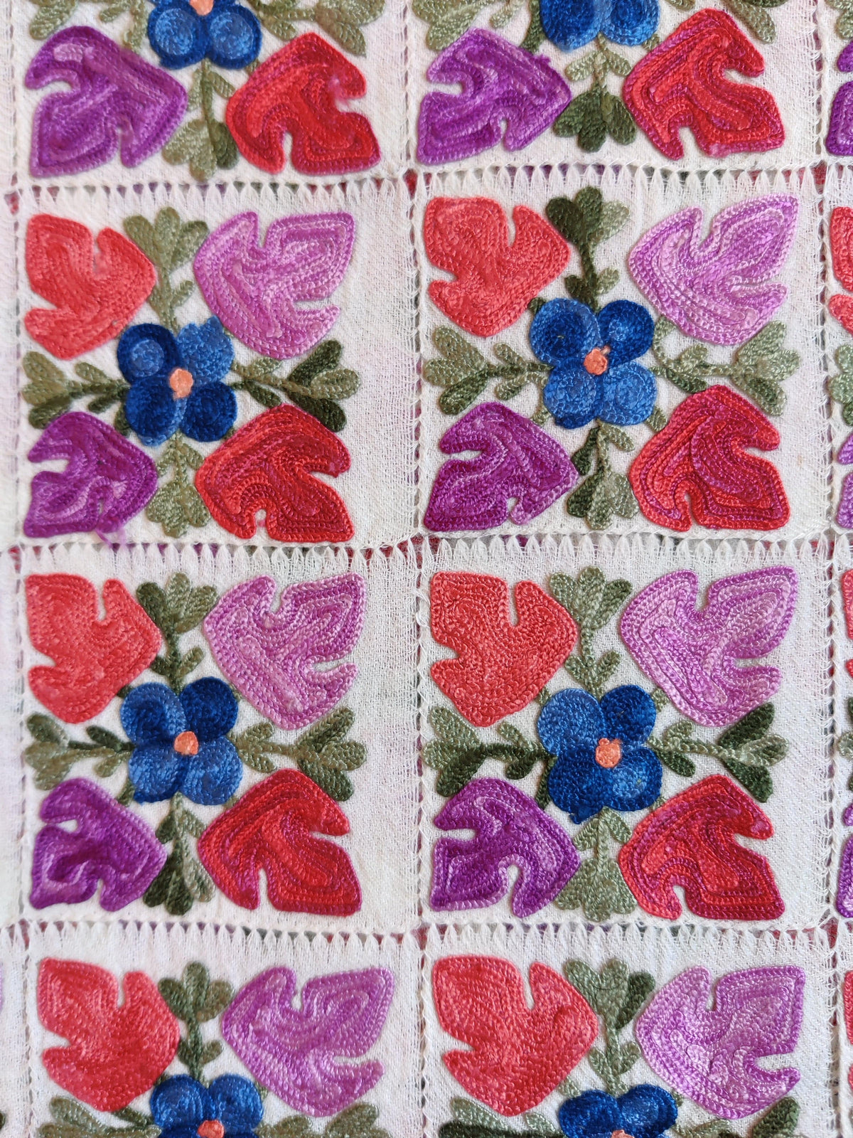 Macro view of blue Aari flowers and pink and purple leaves within white cutwork squares on a Merino wool stole