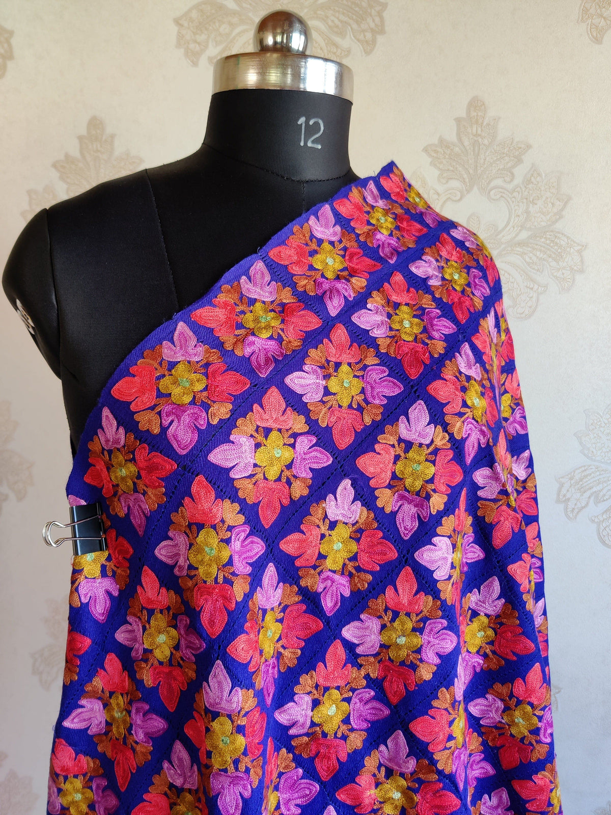 Upper view of a blue merino wool stole on a mannequin, with repeating pink, coral and yellow Aari floral diamonds.