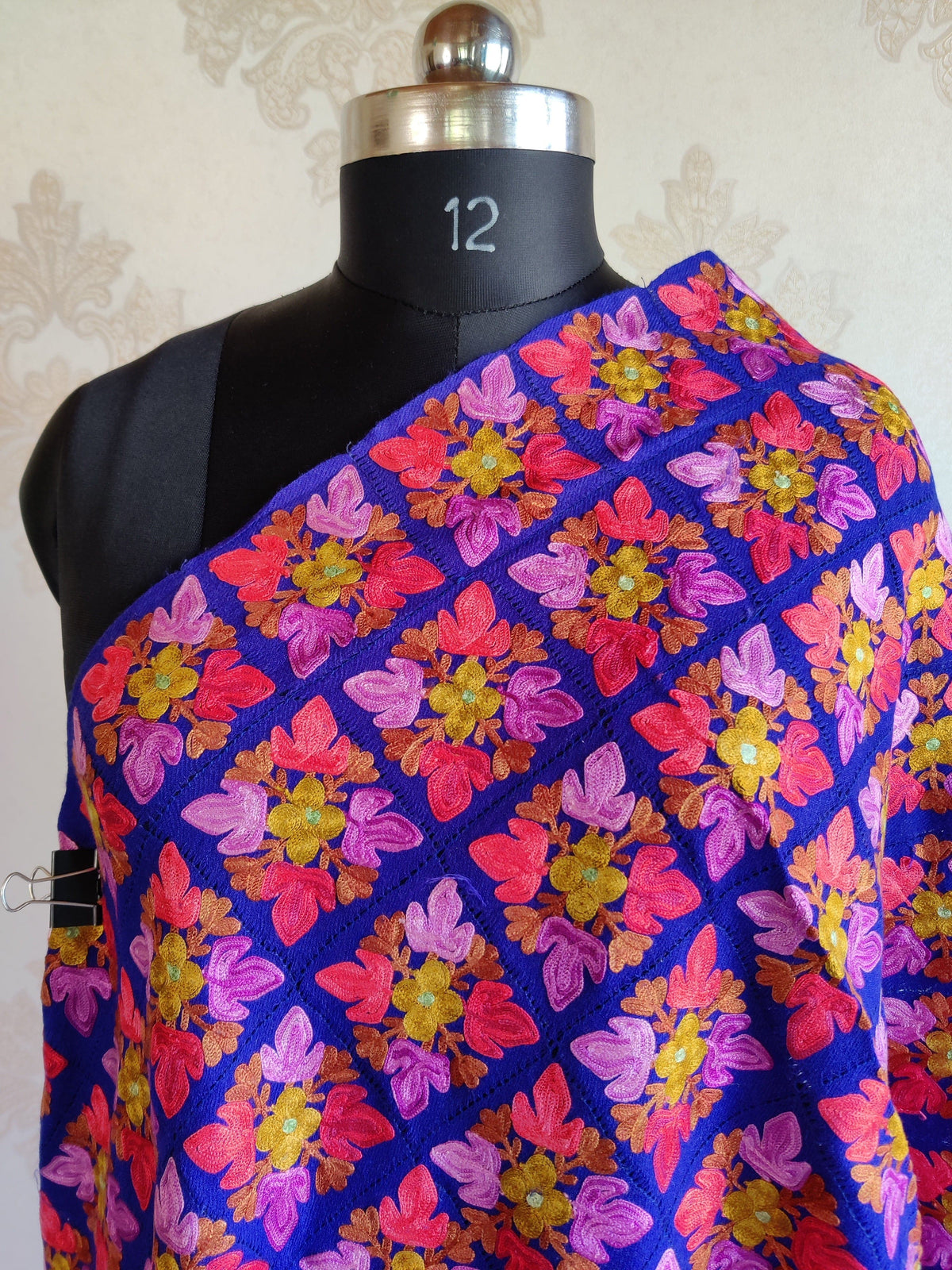 Close-up of a royal-blue merino wool stole across a mannequin's chest, showing pink and coral Aari leaves around yellow flowers.