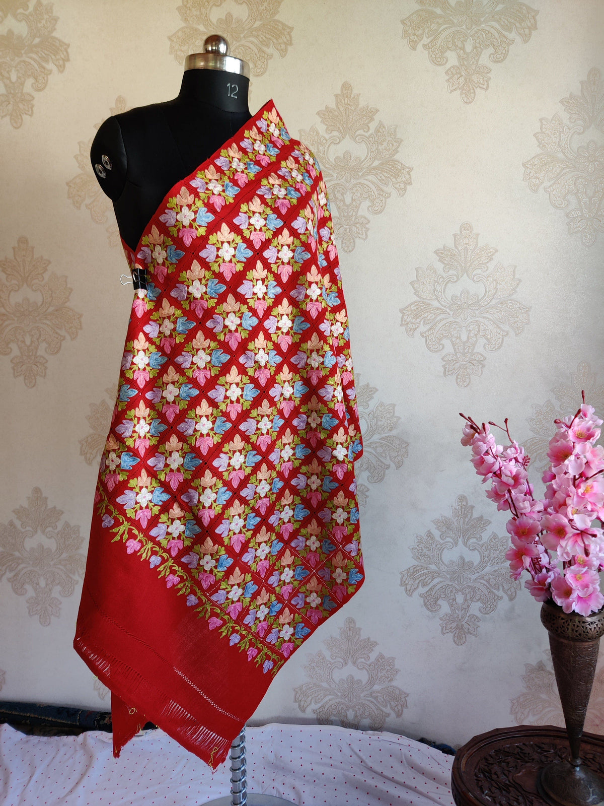 Full view of a red stole on a mannequin, showing multicoloured floral squares, a narrow floral border and plain fringed end.