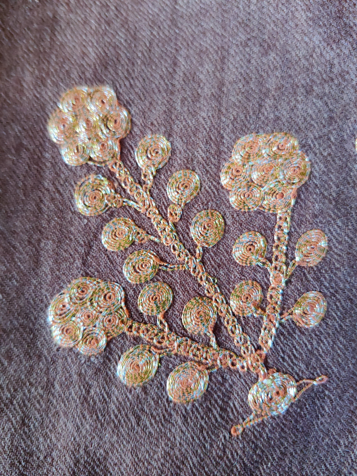 Close-up of a gold-toned Tilla floral sprig with three blooms and round leaves on brown Merino wool