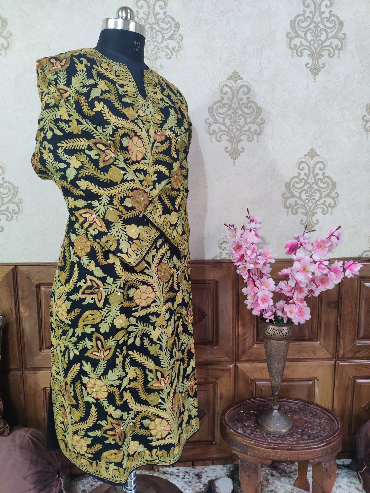Angled full view of a black pheran with crossed sleeves, covered in yellow flowers and fern-like leafy vines.