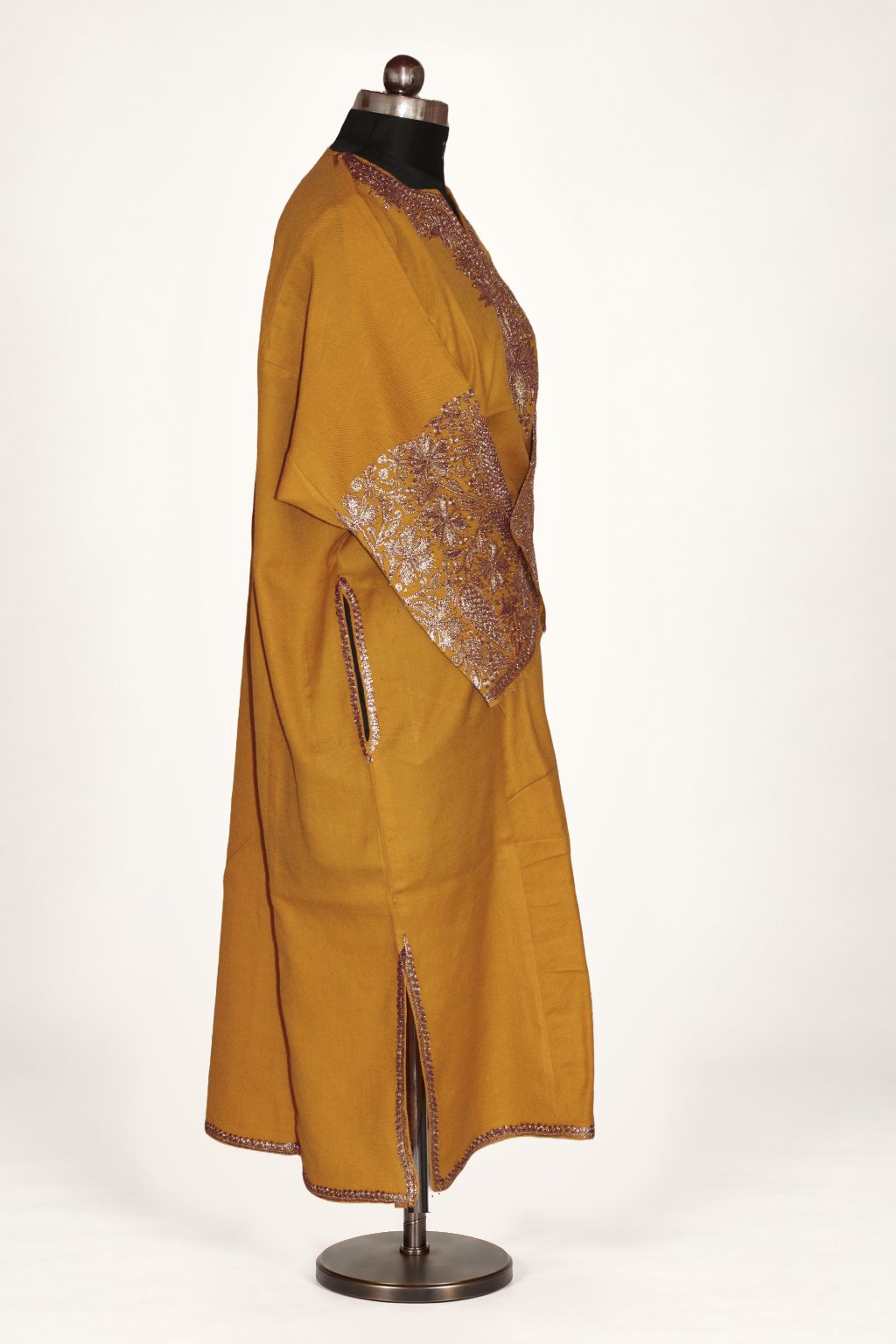 Side view of a mustard pheran on a mannequin, showing pale metallic Tilla floral cuffs and narrow trim at the pocket and side slit.