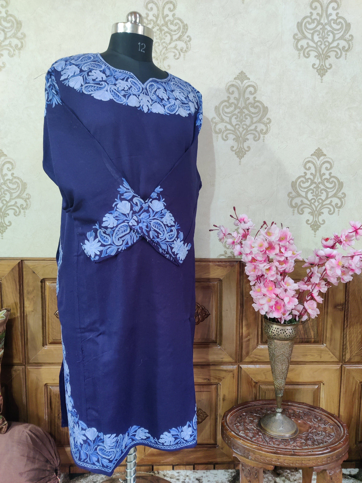 Angled full view of a blue pheran with crossed sleeves and pale blue Aari floral embroidery at the neckline, cuffs and hem