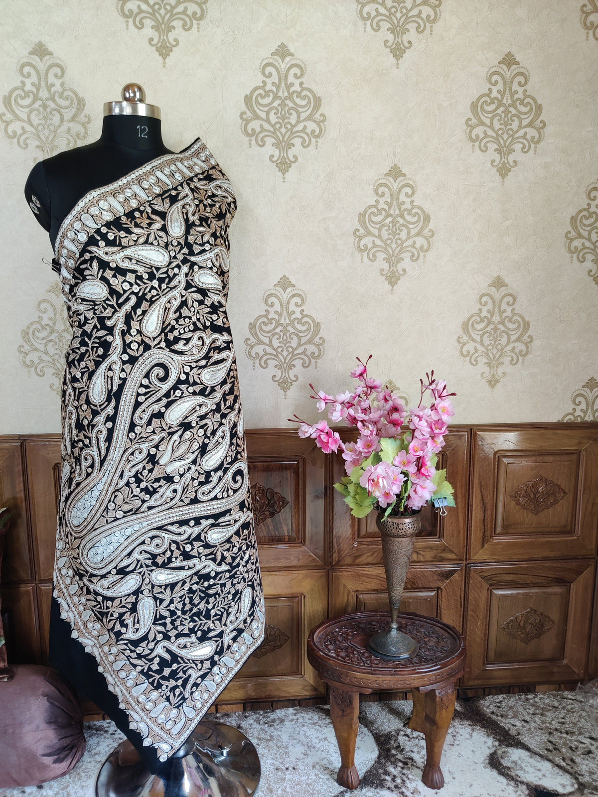 Wide view of the black paisley-embroidered Merino wool shawl wrapped on a mannequin beside a table of pink flowers